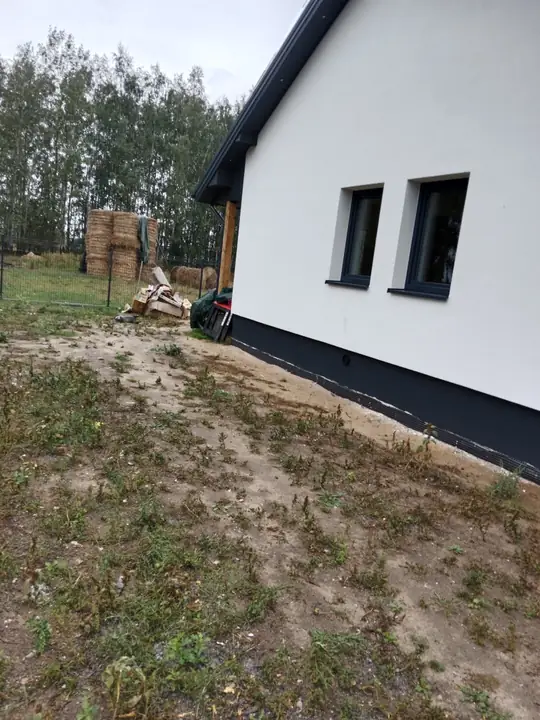 Boczna ściana domu, piasek i trawa przy ogrodzeniu